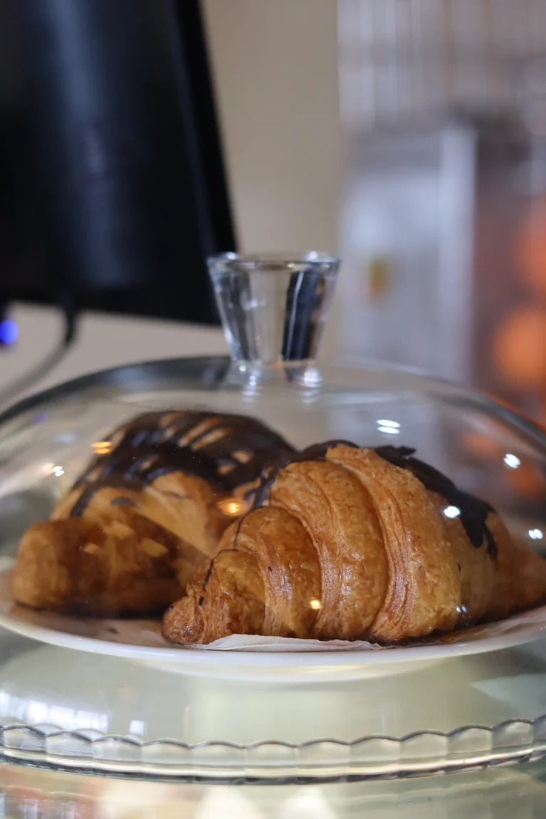 Freshly baked chocolate-drizzled croissants under a glass dome at the counter