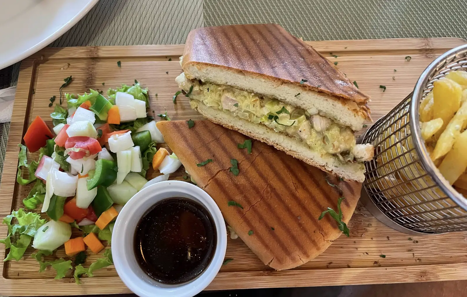 A grilled chicken panini with salad and a basket of fries on a wooden board